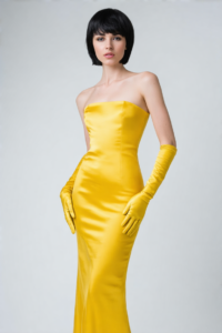 Woman in yellow strapless gown and gloves with short black hair, elegant studio portrait.