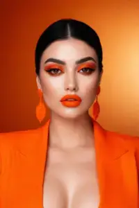 Woman with dark hair and vibrant orange makeup in orange gradient studio, editorial portrait.